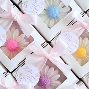 May include: White boxes with daisy-shaped soaps in various colors, including pink, purple, yellow, and blue. Each box is tied with a pink ribbon and features a round sticker with text.