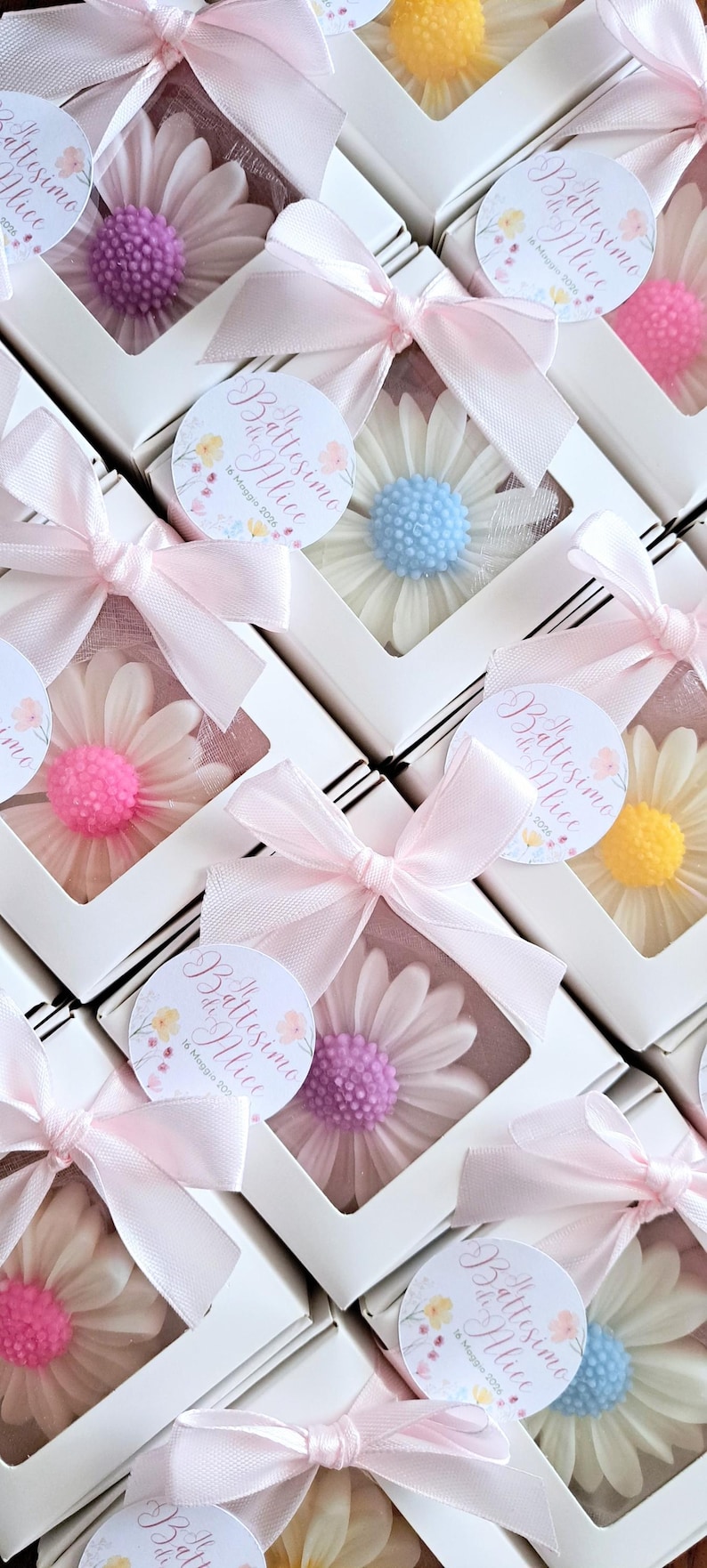 May include: White boxes with daisy-shaped soaps in various colors, including pink, purple, yellow, and blue. Each box is tied with a pink ribbon and features a round sticker with text.