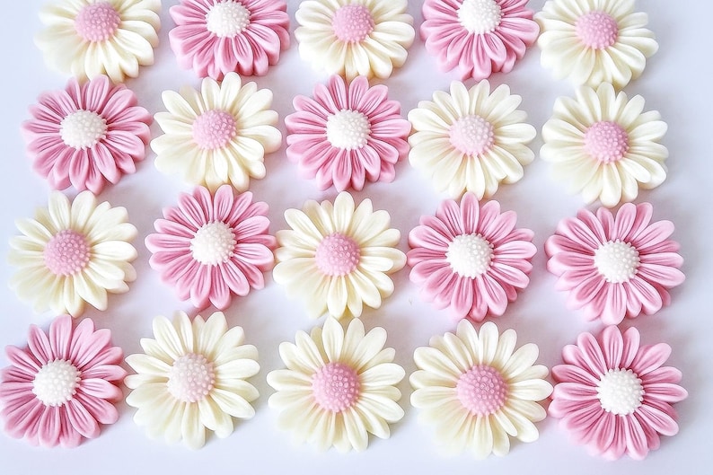 May include: An overhead shot of multiple daisy-shaped decorations. The decorations alternate between pink and off-white petals with a light pink center. The petals are detailed and arranged in a circular pattern, creating a floral design.