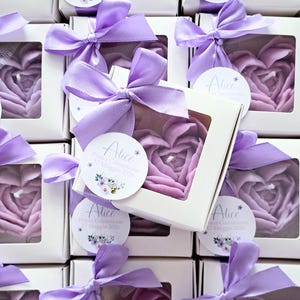 Può includere: Una collezione di scatole regalo quadrate bianche, ognuna contenente una candela viola a forma di cuore. Ogni scatola presenta una finestra trasparente, un fiocco in nastro di raso viola e un'etichetta circolare con il nome Alice e il testo Prima Comunione 17 Maggio 2026.