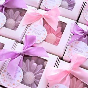 May include: White boxes with clear windows, each containing a flower-shaped soap in shades of pink and purple. The boxes are tied with pink and purple satin ribbons and tags that read "Battesimo di Matilde 31 Maggio 2026".