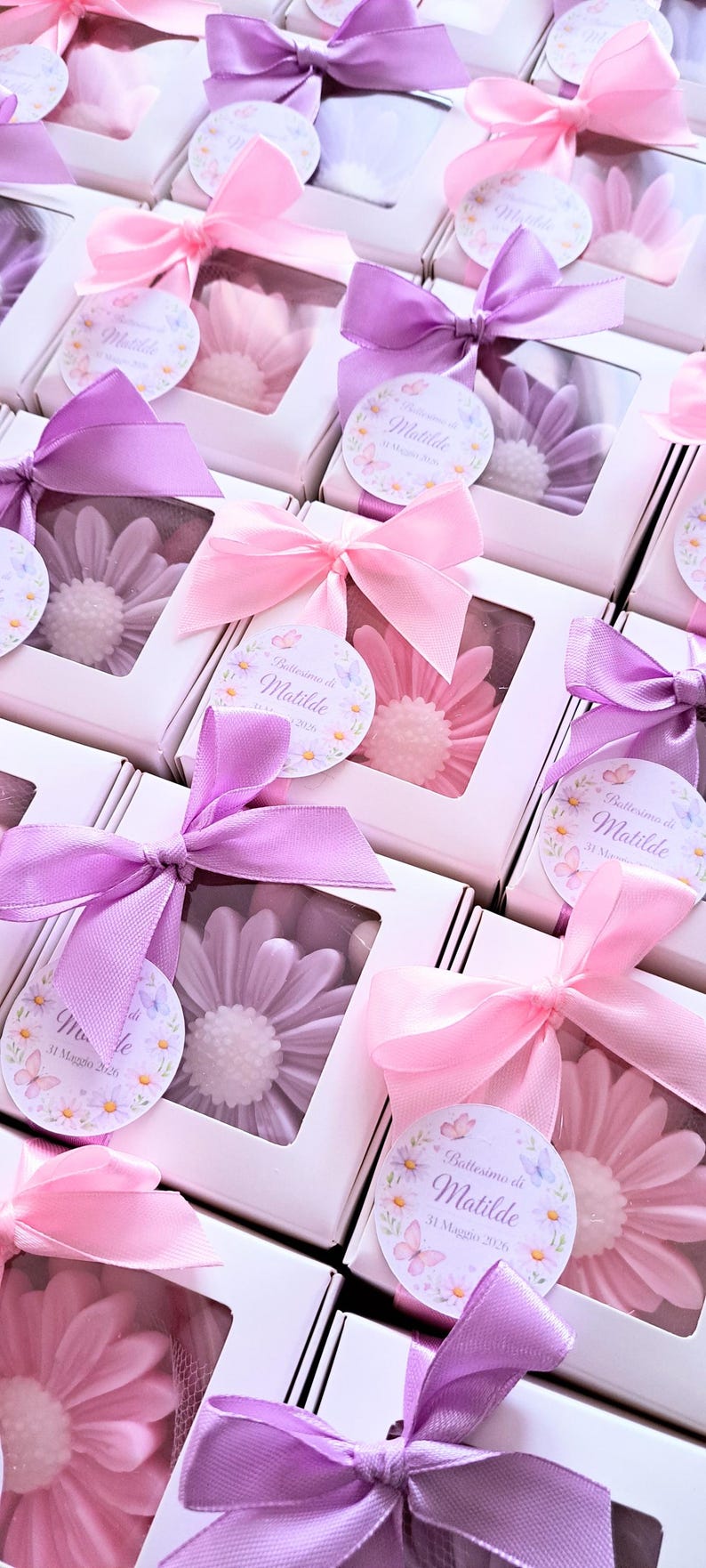 May include: White boxes with clear windows, each containing a flower-shaped soap in shades of pink and purple. The boxes are tied with pink and purple satin ribbons and tags that read "Battesimo di Matilde 31 Maggio 2026".