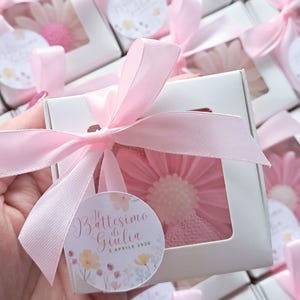 May include: White gift boxes tied with pink ribbons, each containing a pink flower-shaped item. A circular tag with text and floral illustrations is attached to each box. The boxes are arranged in a group.