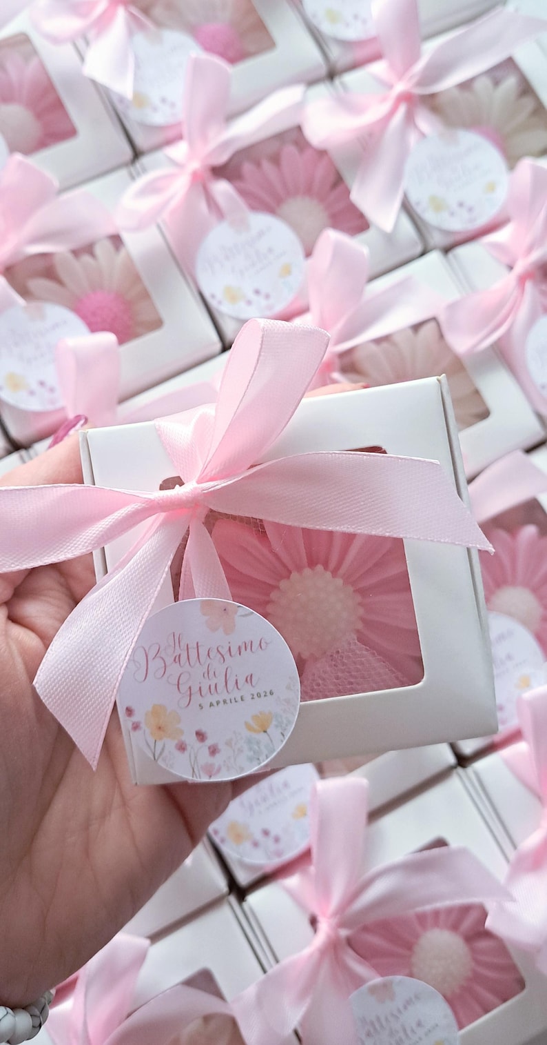 May include: White gift boxes tied with pink ribbons, each containing a pink flower-shaped item. A circular tag with text and floral illustrations is attached to each box. The boxes are arranged in a group.
