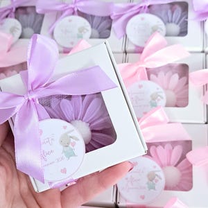 May include: White boxes with a clear window, each containing a flower-shaped item. The boxes are tied with lavender or pink satin ribbons. Each box has a small tag with a bunny illustration and text.