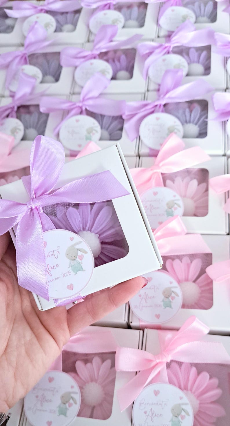May include: White boxes with a clear window, each containing a flower-shaped item. The boxes are tied with lavender or pink satin ribbons. Each box has a small tag with a bunny illustration and text.