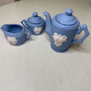 May include: A light blue ceramic tea set featuring a teapot, sugar bowl, and creamer. Each piece has a basketweave pattern and a white floral design. The teapot has a curved spout and handle. The set is displayed on a white surface.