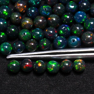 Può includere: Una collezione di perle di opale verde scuro e nero con riflessi iridescenti di colore. Le perle sono disposte in un mucchio sciolto su una superficie nera, con una pinzetta sopra.