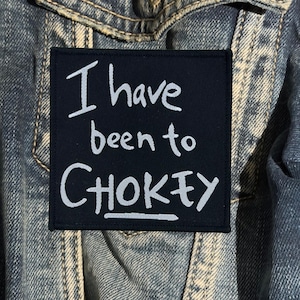 May include: A square, dark navy blue embroidered patch featuring the light blue text "I have been to CHOKEY" with a horizontal line underneath the word CHOKEY. The patch has a stitched border and is placed on a blue denim fabric background.