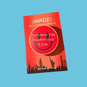 Puede incluir: Pin de madera rojo con una cara sonriente y el texto "You're Never Fully Dressed Without A Smile". El pin es parte de la colección de teatro musical "Annie", con un diseño del horizonte de Nueva York.