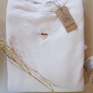 May include: A pale pink sweatshirt with a small embroidered image of a steaming cup of coffee. The sweatshirt has a brown tag attached with the text "Little Things" on it.