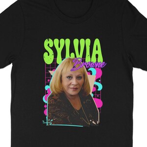 May include: Black t-shirt with a retro design featuring the name "Sylvia Browne" and a portrait of a woman with blonde hair. The graphic includes neon green, pink, and blue elements. The t-shirt is a classic crew neck style.
