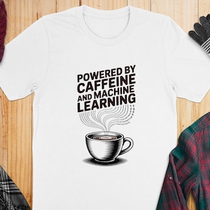 Peut inclure: T-shirt blanc avec le texte "POWERED BY CAFFEINE AND MACHINE LEARNING" au-dessus d'un dessin de tasse à café. Le motif représente une tasse à café avec de la vapeur qui s'élève et du code binaire. Le t-shirt est présenté sur une surface en bois.