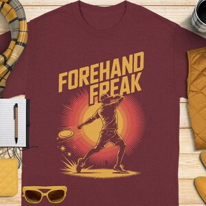 Peut inclure: T-shirt bordeaux avec l'inscription jaune « FOREHAND FREAK » et un graphisme d'une personne jouant au disc golf. Le design comprend un effet de soleil derrière la silhouette et un disque volant.