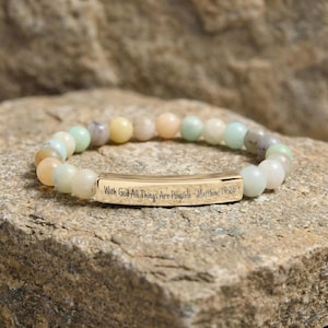 May include: A bracelet with pastel-coloured beads and a gold-coloured bar. The bar is inscribed with the words "With God All Things Are Possible - Matthew 19:26". The bracelet is displayed on a stone surface. The beads are approximately 0.8 cm in diameter.