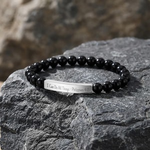May include: A black beaded bracelet featuring a silver bar engraved with the words "I Can Do All Things - Philippians 4:13". The bracelet is displayed on a grey stone surface.