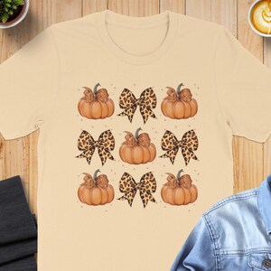 Könnte beinhalten: Ein beiges T-Shirt mit einem Muster aus orangefarbenen Kürbissen und braunen und schwarzen Leoparden-Schleifen.