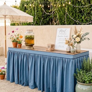 May include: A rectangular wooden bar table draped with a pleated dusty blue fabric skirt. The table holds a glass beverage dispenser, a framed sign reading M&C Wedding Bar Signature Drinks, small floral arrangements, and a stack of cards.