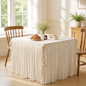 May include: A square table covered with a floor-length, off-white pleated tablecloth. The fabric features vertical pleats and is secured at each corner with matching fabric ties. The table surface holds a glass vase with flowers, two croissants on a plate, a small jar, a bowl, and a mug.