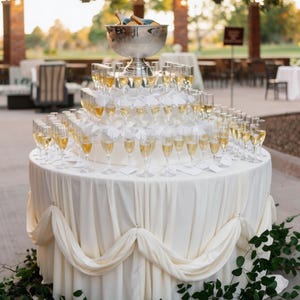 May include: A tiered champagne display at an outdoor event. The table is covered in a cream-colored cloth and adorned with greenery. A large silver bowl holds champagne bottles, and the glasses are arranged in a pyramid.