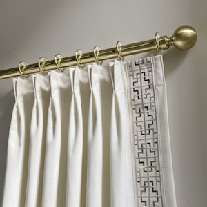 Puede incluir: Cortinas de lino blanco con un borde de patr&oacute;n geom&eacute;trico que cuelgan de una barra de cortina dorada con terminales.