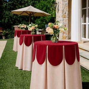 May include: Outdoor event tables with burgundy scalloped valances and beige skirts. Each table is topped with a vase of flowers. The tables are arranged on a green lawn, with a beige umbrella in the background.