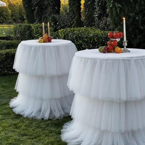 May include: Two round tables with white tiered tulle skirts are set on a green lawn. Each table is topped with a white tablecloth and decorated with fruit and candles. The background features manicured hedges and trees.