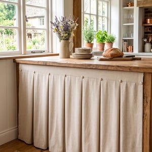 May include: A kitchen island featuring a beige fabric skirt with box pleats and matching fabric-covered buttons along the top edge. The island has a natural wood countertop holding a ceramic pitcher with dried flowers and a stack of plates.