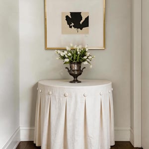 Peut inclure: Une table ronde crème avec une jupe plissée et des boutons décoratifs. Un vase argenté contient un bouquet de tulipes blanches. Au-dessus, une œuvre d'art abstraite encadrée avec des éléments noirs et beiges est accrochée au mur.