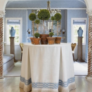 May include: A round white tablecloth with a blue geometric border and fringe. The table holds potted topiary trees. The room has blue and white checkered wallpaper, blue window treatments, and decorative urns on pedestals.