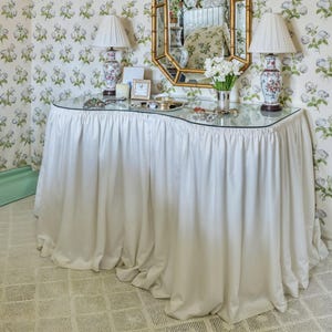 Puede incluir: Una mesa de tocador blanca con falda y tapa de cristal, adornada con objetos decorativos, incluyendo lámparas, un espejo y un jarrón con flores. La mesa se encuentra frente a un fondo de papel pintado floral, creando una estética elegante y clásica.