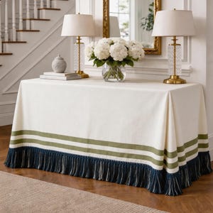 Peut inclure: Une nappe rectangulaire blanc cassé ornée de deux bandes horizontales vert olive près du bord inférieur, terminée par une frange bleu marine profonde qui tombe jusqu'au sol, recouvrant les côtés d'une table console.