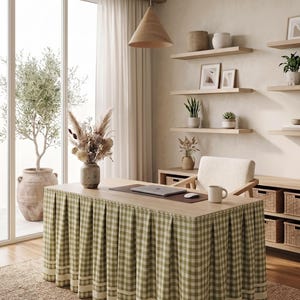 May include: A rectangular desk featuring a light wood top and a floor-length, pleated skirt in a sage green and white gingham check pattern. The desk surface holds a laptop, a computer mouse, a ceramic mug, and a vase filled with dried flowers.