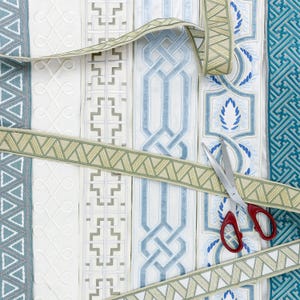 May include: An assortment of decorative ribbons in various patterns and colors, including blue, white, and green. A pair of scissors with red handles is positioned on top of the ribbons, suggesting a crafting or design context.