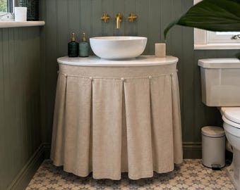 May include: A bathroom vanity featuring a white vessel sink and gold faucet, enclosed by a beige fabric skirt with box pleats and small circular buttons along the top edge. The vanity is set against a sage green paneled wall with a round mirror.