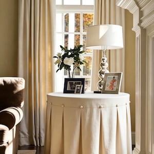Pode incluir: Uma mesa lateral redonda com uma saia plissada creme e botões decorativos. Uma lâmpada prateada com uma sombra branca, duas fotos emolduradas e um vaso de flores brancas estão em cima. A mesa está numa sala com cortinas creme.