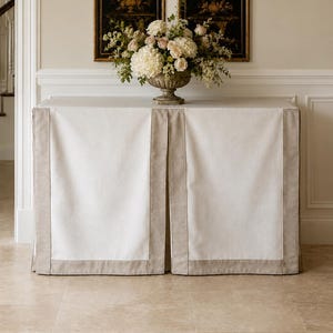 May include: A rectangular console table draped in a floor-length white fabric cover with wide, light beige border panels along the edges. A decorative urn-shaped vase filled with white and pale pink flowers sits centered on the table surface.