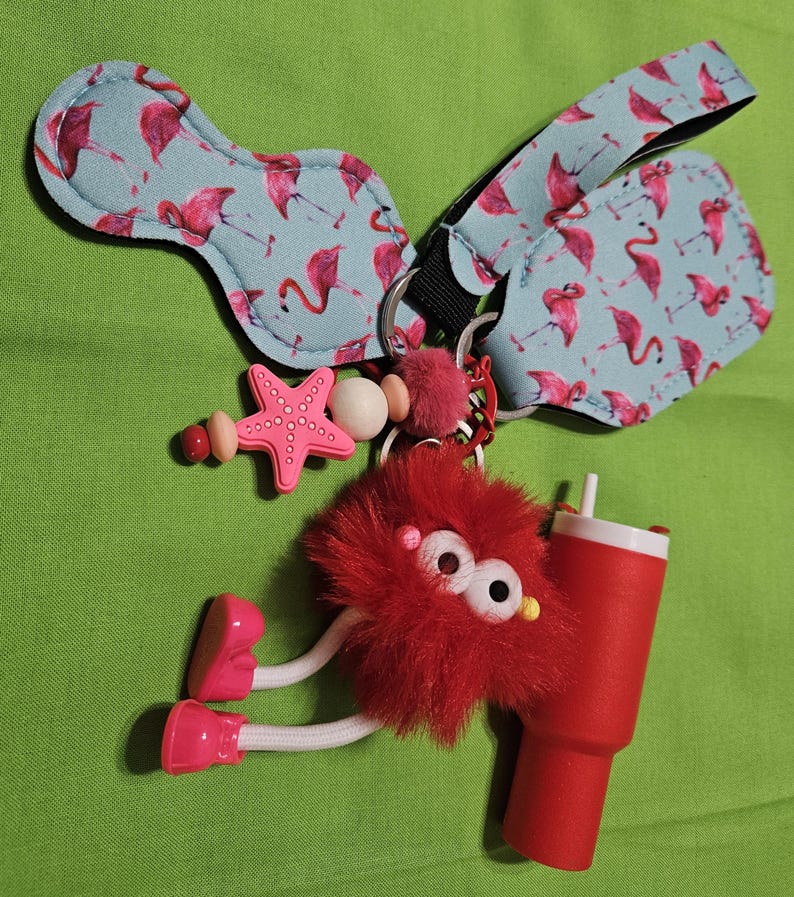 May include: A collection of keychains and accessories featuring a flamingo pattern, a red fuzzy character with a drink, and a pink starfish. The flamingo pattern is on a light blue background. The accessories are attached to a keyring.