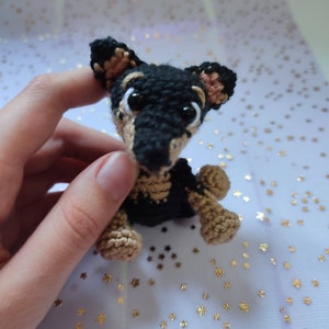 Puede incluir: Un pequeño juguete de perro de crochet con pelo negro y marrón, ojos marrones y nariz negra.