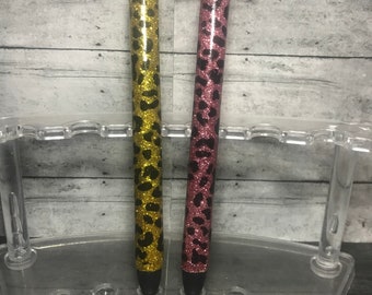 May include: Two glitter pens with black leopard print designs. One pen is gold glitter and the other is pink glitter.