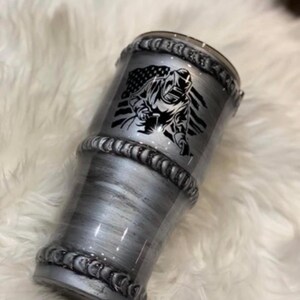 May include: A silver-toned stainless steel tumbler featuring a brushed metallic finish and raised, textured bands resembling industrial metal welds. The design includes a black graphic of a welder wearing a helmet in front of an American flag.