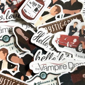 Puede incluir: Una colección de pegatinas troqueladas con imágenes y texto de "The Vampire Diaries". Las pegatinas incluyen imágenes de personajes, un coche rojo y frases como "Hello, love" y "Welcome to Mystic Falls."