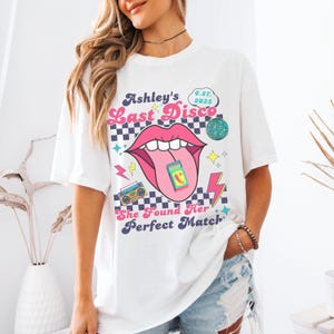 May include: White t-shirt with a graphic design featuring a pink tongue sticking out of a mouth with teeth. The text "Ashley's Last Disco 6.27. 2025" is above the mouth. The text "She Found Her Perfect Match" is below the mouth.