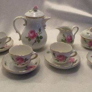 Puede incluir: Un juego de té de porcelana blanca que incluye una tetera con tapa, una lechera, un azucarero con tapa y cuatro tazas con sus platos a juego. Cada pieza está decorada con rosas rosadas, hojas verdes y bordes con detalles dorados.