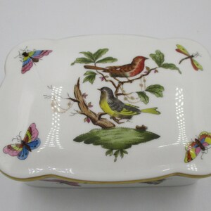 Puede incluir: Caja de baratijas de porcelana blanca con borde festoneado y ribete dorado. La tapa presenta una ilustración detallada de dos pájaros posados en una rama, rodeados de mariposas y una libélula. El objeto es decorativo.