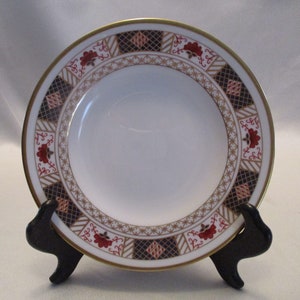 Peut inclure: Une assiette en céramique blanche avec un bord doré et une bordure à motifs rouge, bleu et or. L'assiette est sur un support noir.