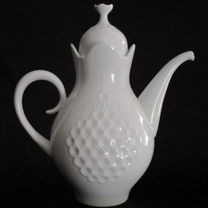 May include: A white ceramic teapot with a lid and a curved handle. The teapot features a textured, honeycomb-like pattern on its body. The spout is long and curved, and the lid has a floral-shaped finial. The background is black.