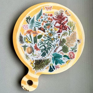 May include: Round, yellow cutting board with a handle, decorated with botanical stickers of flowers, butterflies, and foliage. The board has a yellow and white striped border and a hole in the handle for hanging.