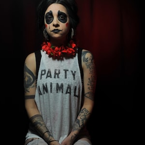May include: A person with panda-style makeup, wearing a gray tank top with the words "PARTY ANIMAL" printed on it. They are adorned with a red flower necklace and have multiple tattoos on their arms and legs. The background is dark.
