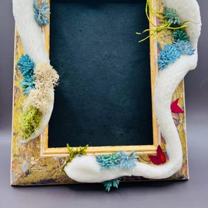 May include: A decorative picture frame with a gold and distressed finish. The frame is adorned with white, textured accents and clusters of blue, green, and red floral elements. The frame's interior is a dark, matte black.